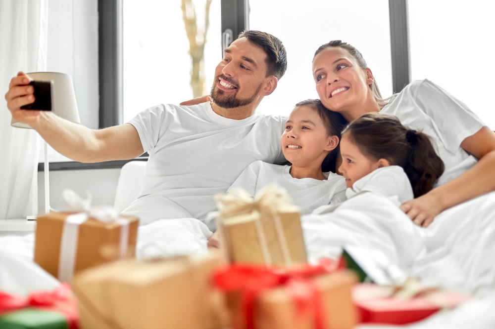 family, winter holidays and people concept - happy mother, father and two daughters with christmas gifts taking selfie in bed at home. family with christmas gifts taking selfie in bed