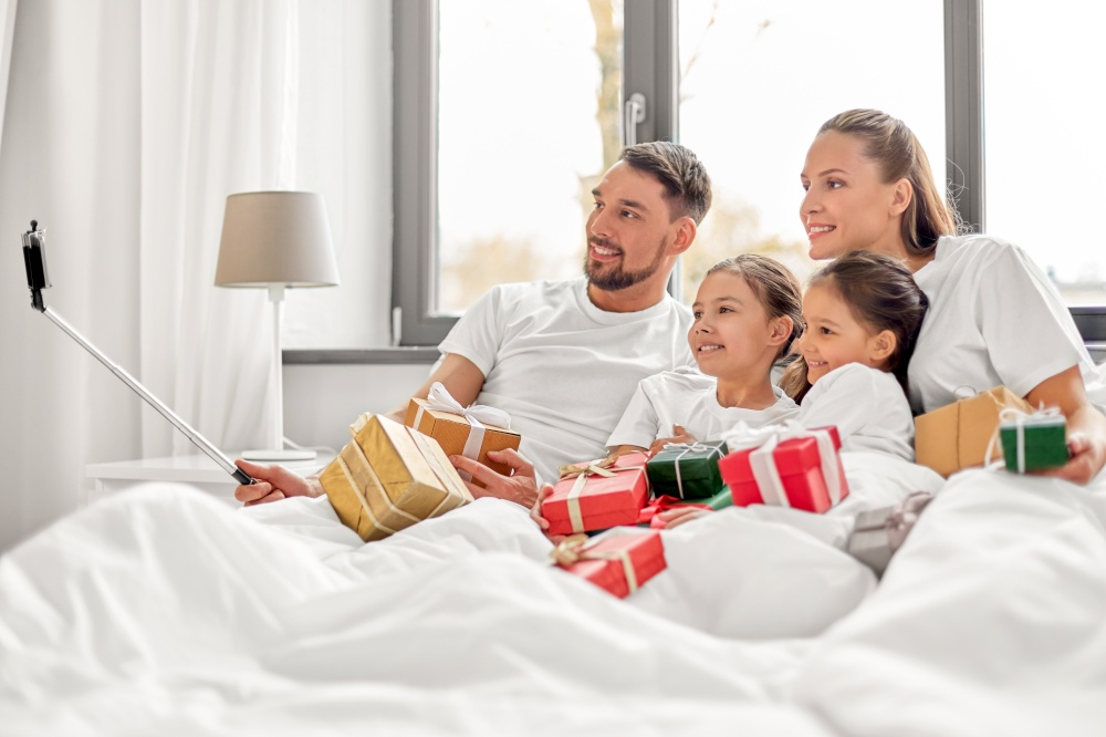 family, winter holidays and people concept - happy mother, father and two daughters with christmas gifts taking selfie in bed at home. family with christmas gifts taking selfie in bed