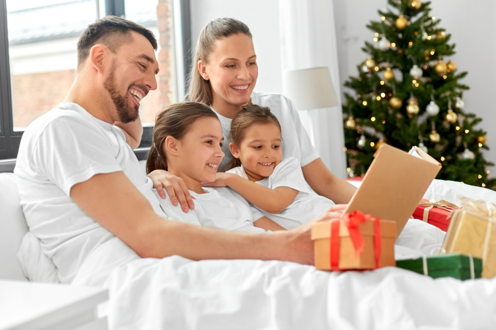 family, winter holidays and people concept - happy mother, father and two daughters in pajamas with christmas gifts reading book in bed at home. happy family reading book in bed on christmas