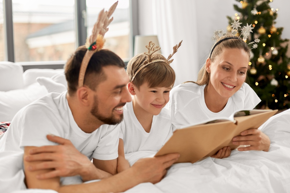 family, winter holidays and people concept - happy mother, father and little son in pajamas reading book in bed in christmas morning. family with book in pajamas in bed on christmas