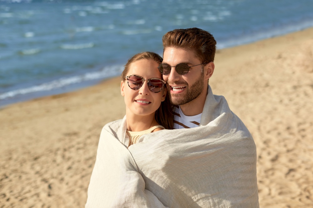 leisure, relationships and people concept - happy couple in sunglasses covered with blanket hugging on summer beach. happy couple covered with blanket hugging on beach