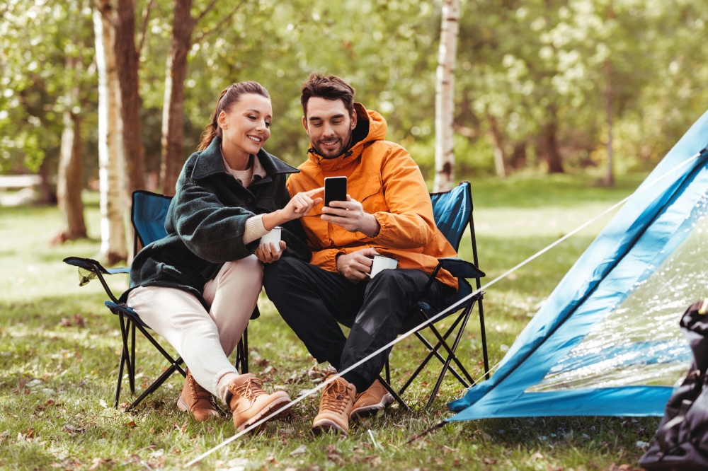 camping, tourism and travel concept - happy couple with smartphone drinking tea at campsite. couple with smartphone drinking tea at campsite