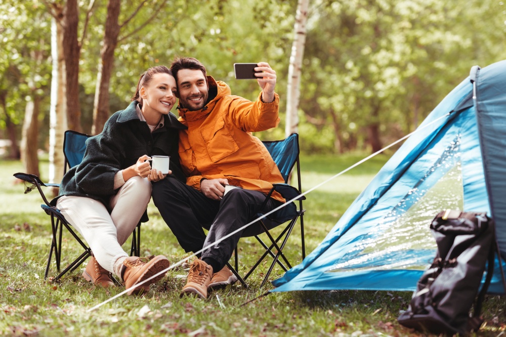 camping, tourism and travel concept - happy couple with smartphone drinking tea and taking selfie at tent camp. couple drinking tea and taking selfie at tent camp