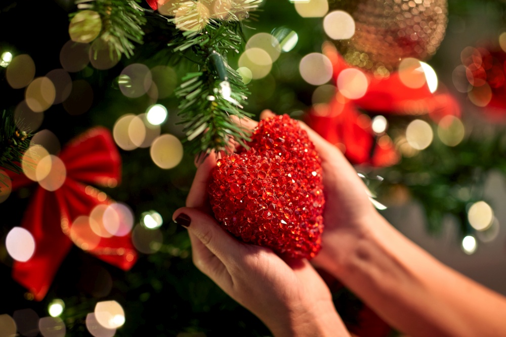 holidays, celebration and people concept - woman hands decorating christmas tree with red heart. hands decorating christmas tree with red heart