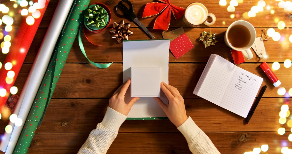 holidays, new year and christmas concept - hands wrapping gift box into green paper on wooden table over festive lights. hands wrapping christmas gift into paper at home