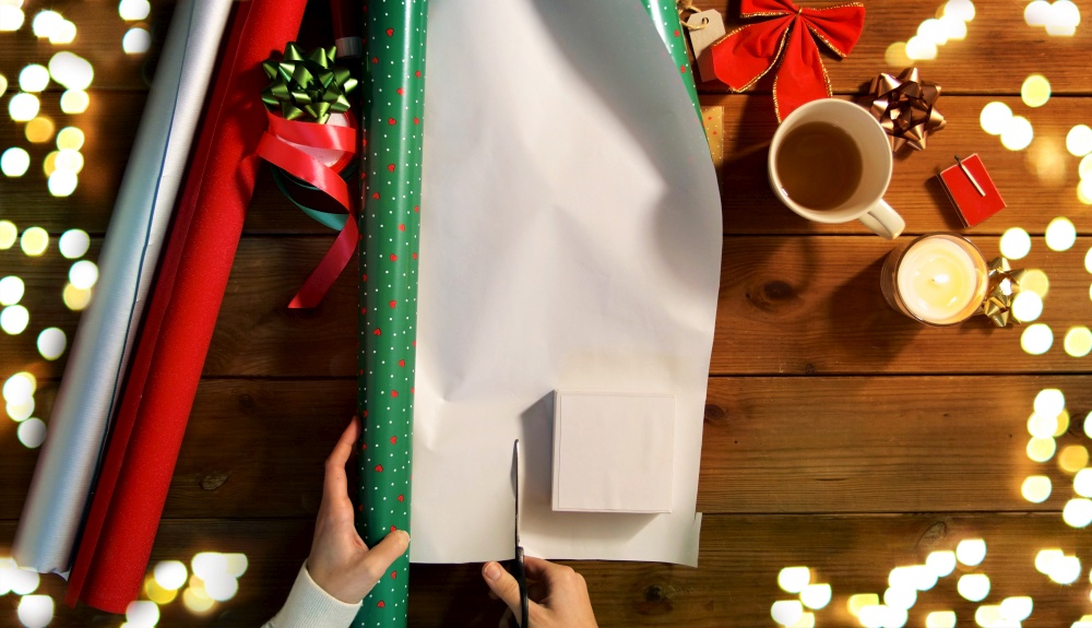 holidays, new year and christmas concept - hands wrapping gift box into green paper on wooden table over festive lights. hands wrapping christmas gift into paper at home