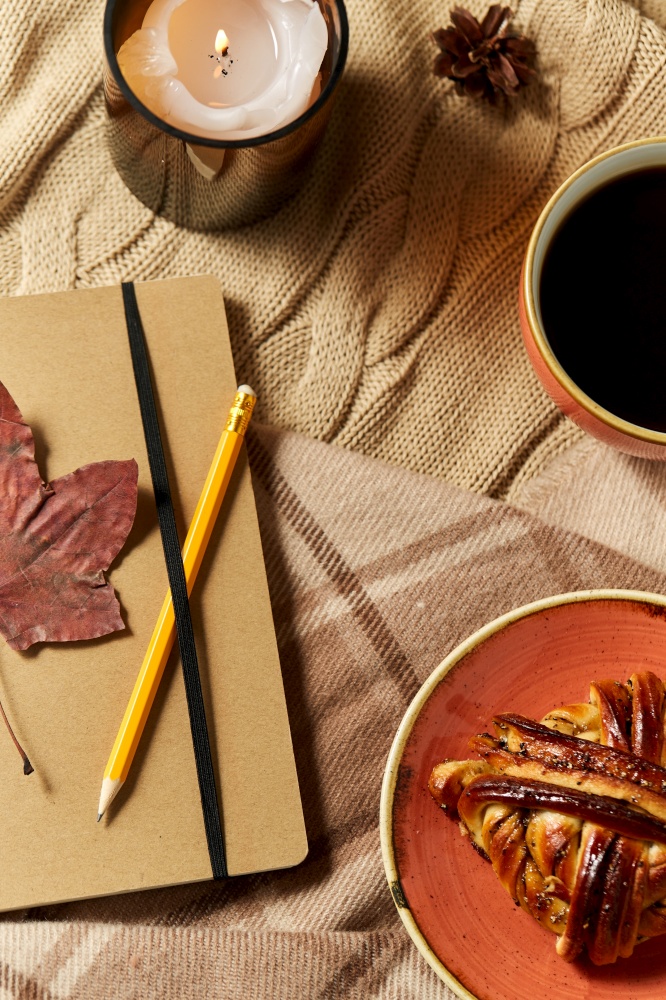 autumn, season and leisure concept - notebook or diary with pencil, cup of coffee, cinnamon bun and candle on warm blankets at home. diary, cinnamon bun, coffee and candle in autumn