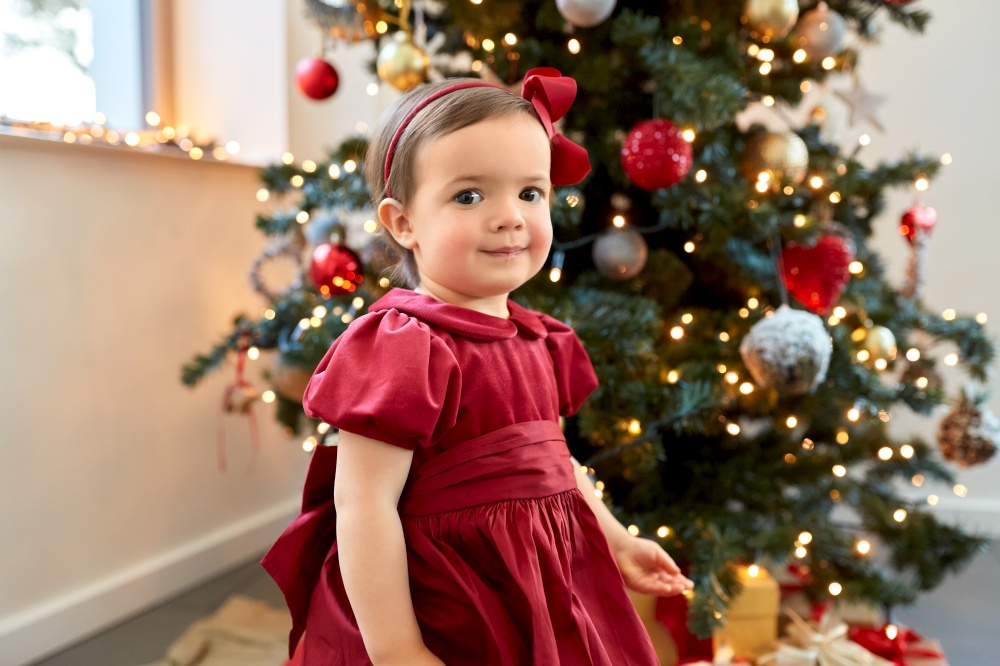 winter holidays and childhood concept - lovely baby girl in red dress over christmas tree at home. lovely baby girl over christmas tree at home