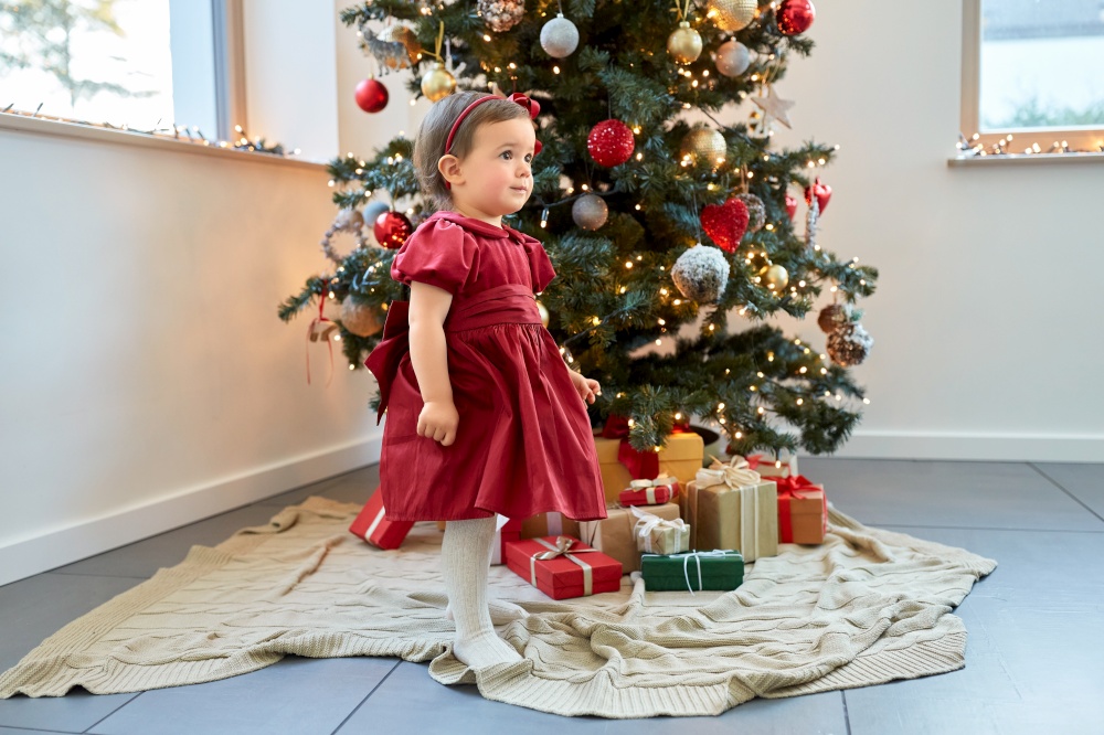 winter holidays and childhood concept - lovely baby girl in red dress over christmas tree at home. lovely baby girl over christmas tree at home