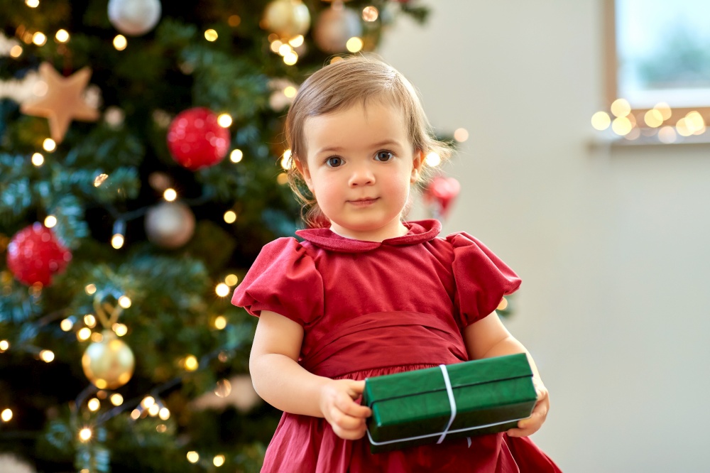 christmas, holidays and childhood concept - happy baby girl in red dress holding green gift at home. happy baby girl with christmas gift at home