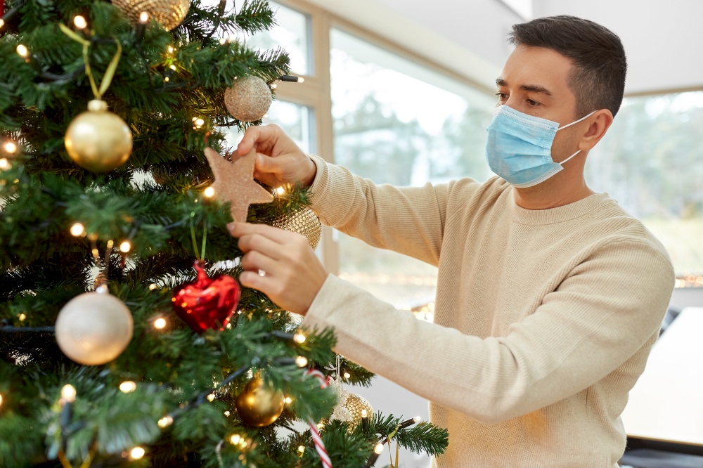 winter holidays, health and people concept - middle aged man in medical mask decorating christmas tree at home. man in mask decorating christmas tree at home