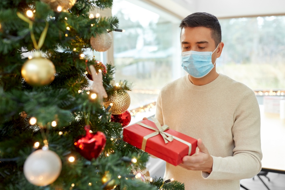 winter holidays, health and people concept - middle aged man in medical mask with gift box at christmas tree at home. man in mask with gift at christmas tree at home
