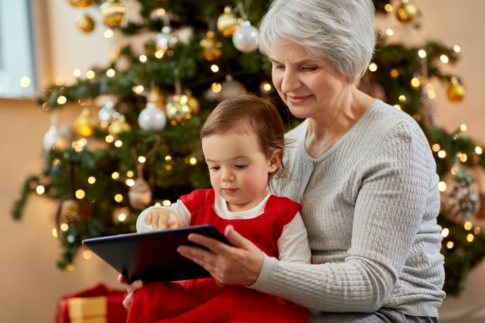 winter holidays and family concept - happy grandmother and baby granddaughter with tablet pc computer sitting near christmas tree at home. grandmother and baby girl with christmas gifts