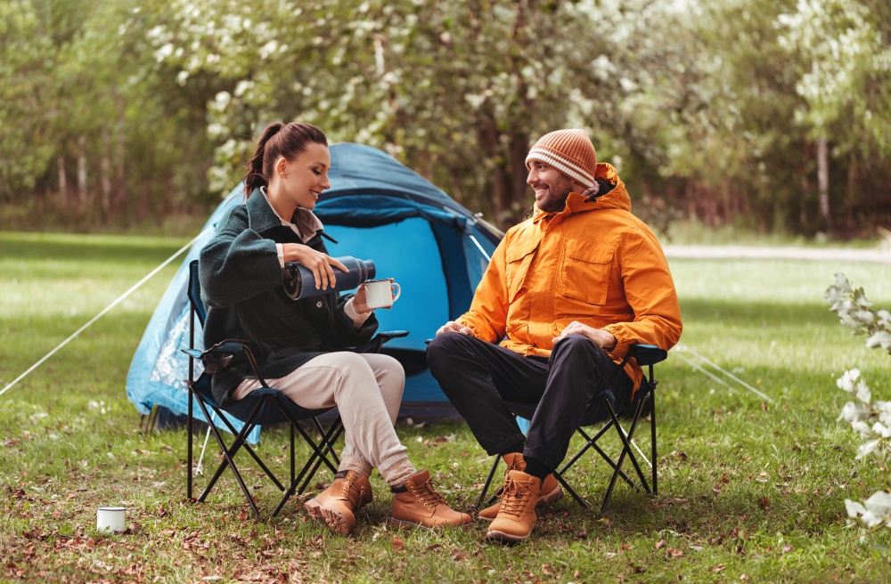 camping, tourism and travel concept - happy couple with thermos drinking tea at tent camp. happy couple drinking tea at tent camp