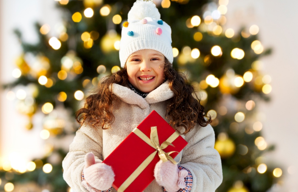 holidays, christmas and people concept - happy little girl with red gift box at winter park over christmas tree lights on background. happy little girl with gift over christmas tree