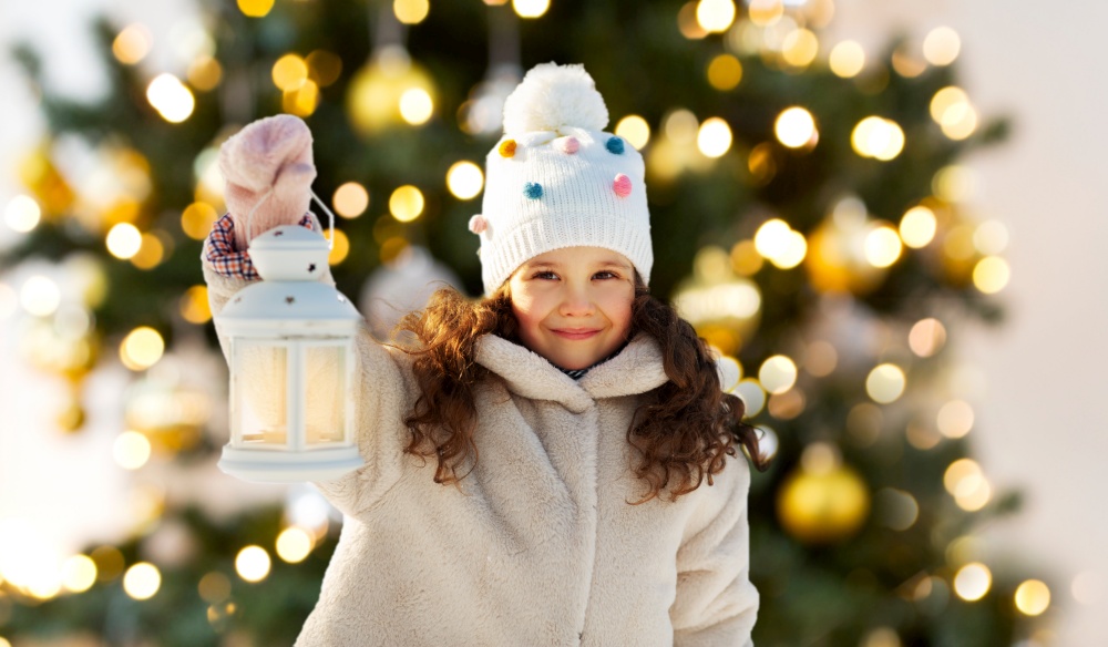 childhood, christmas and season concept - portrait of happy little girl in winter clothes with lantern outdoors at park over christmas tree lights on background. happy little girl with lantern over christmas tree