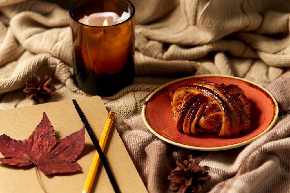 autumn, season and leisure concept - notebook or diary with pencil, cinnamon bun and candle on warm blankets at home. diary, pencil, cinnamon bun and candle in autumn