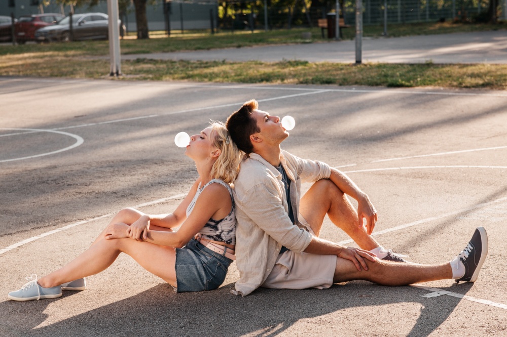 summer holidays and people concept - happy young couple sitting back to back and blowing bubble gums on basketball playground. couple sitting on ground and blowing bubble gums