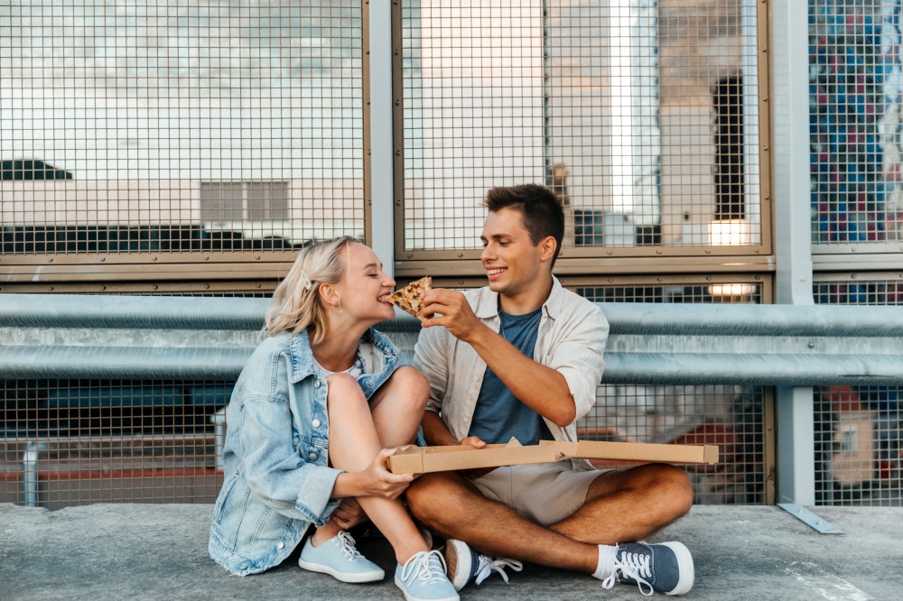 summer holidays, food and people concept - happy young couple eating takeaway pizza on city roof top parking. happy couple eating pizza on city roof top parking