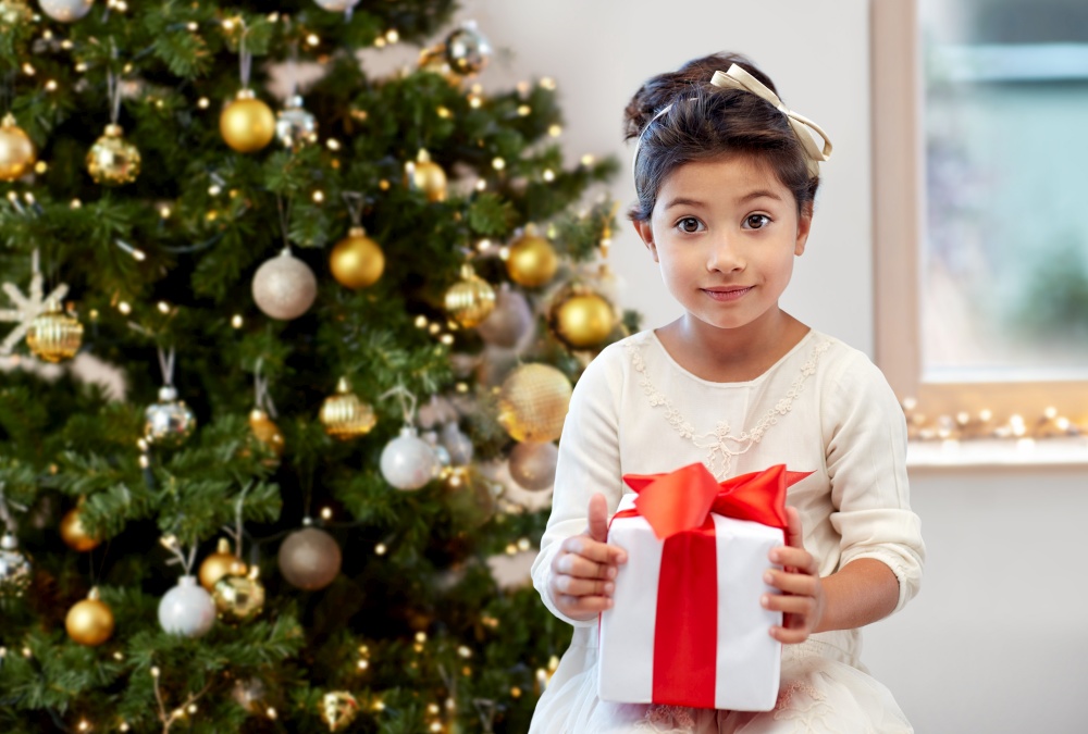 winter holidays, childhood and people concept - smiling little girl with gift box over christmas tree on background. happy little girl with gift over christmas tree