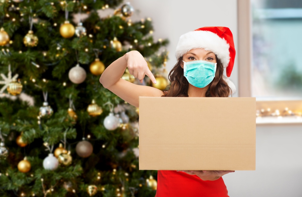 winter holidays, delivery and people concept - woman in mask and santa helper hat with parcel box over christmas tree on background. woman in mask with parcel box on christmas