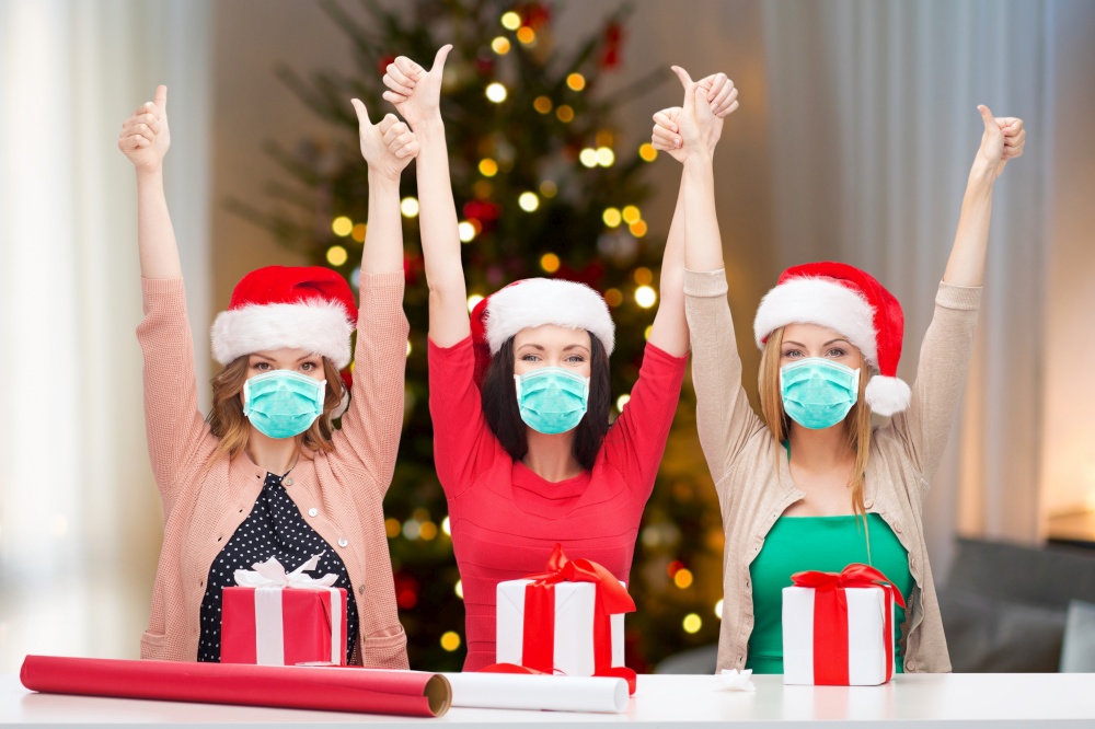 winter holidays and people concept - three happy women in masks packing gifts and showing thumbs up at home over christmas tree on background. women in masks packing christmas gifts at home