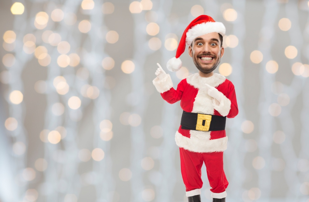 emotion, expression and winter holidays concept - happy smiling young man in santa claus costume showing something over christmas lights background (funny cartoon style character with big head). smiling man in santa costume over christmas tree