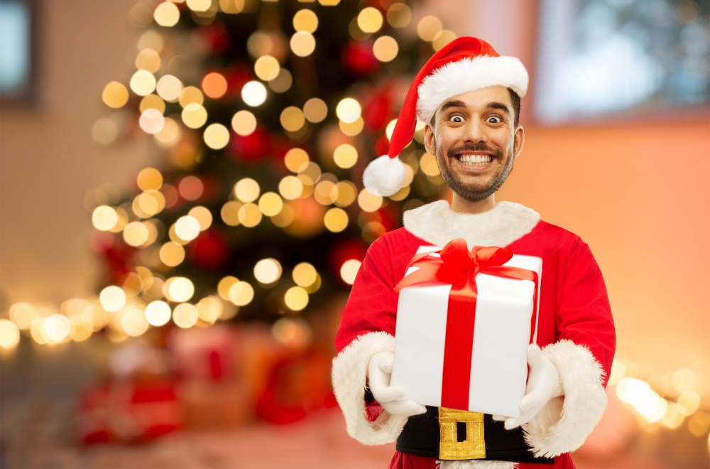 emotion, expression and winter holiday concept - happy smiling young man in santa claus costume over christmas tree lights background (funny cartoon style character with big head). smiling man in santa costume over christmas tree
