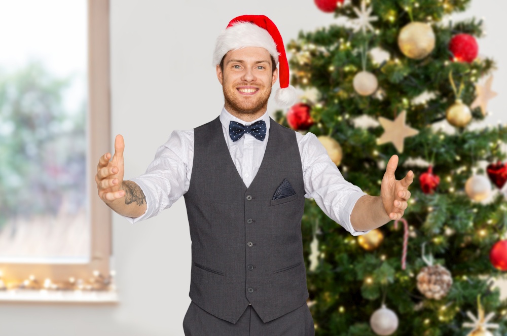 winter holidays and people concept - happy man in santa hat holding something imaginary over christmas tree background. happy man in santa hat holding something imaginary