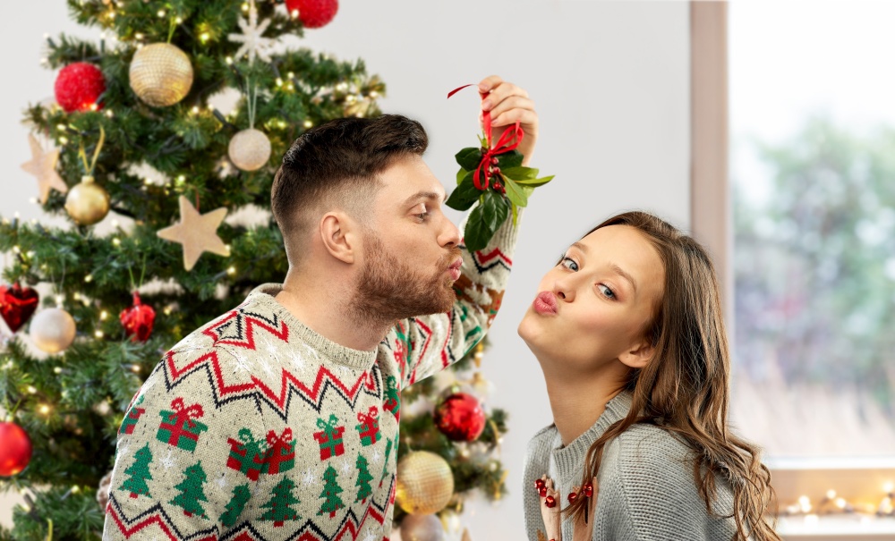 winter holidays, people and traditions concept - happy couple in ugly sweaters kissing under the mistletoe over christmas tree on background. couple kissing under the mistletoe on christmas