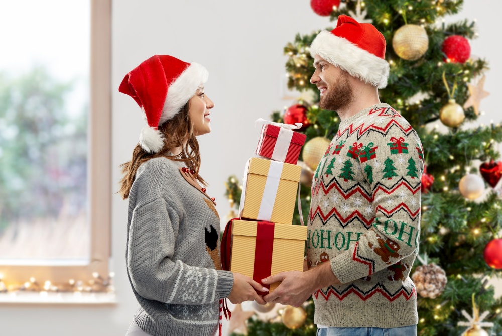 people and holidays concept - happy couple in santa hats with gifts at ugly sweater party over christmas tree on background. happy couple in christmas sweaters with gifts