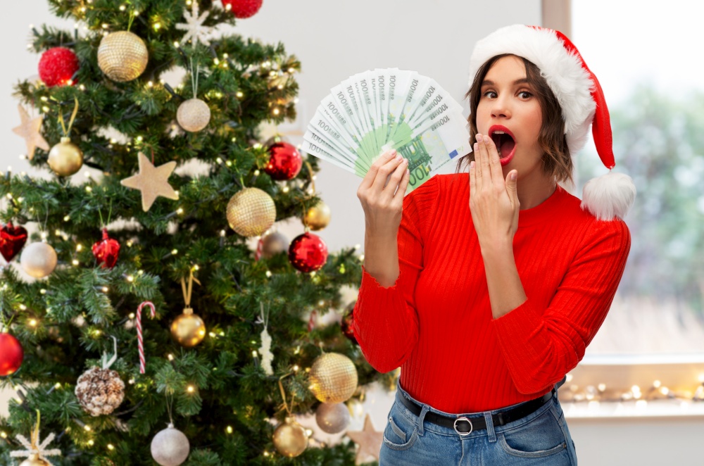winter holidays and finance concept - surprised young woman in santa helper hat holding euro money banknotes over christmas tree on background. surprised woman in santa hat with money