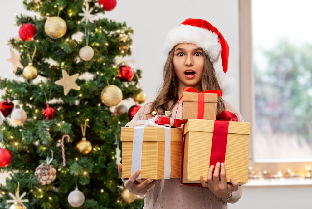 holidays and people concept - shocked teenage girl in santa helper hat holding gift box over christmas tree on background. teenage girl in santa hat with christmas gift