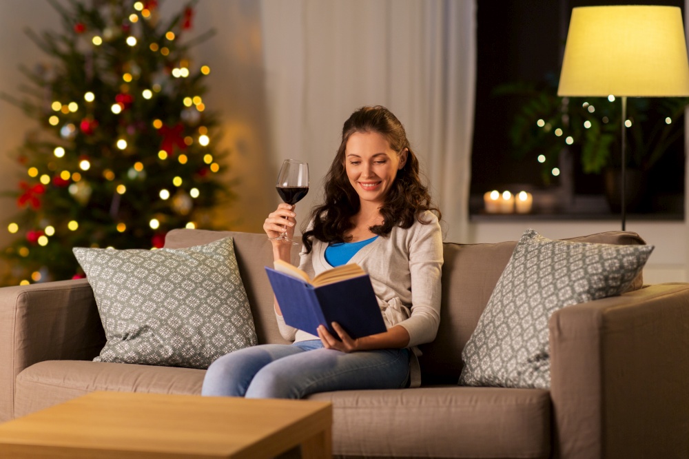 leisure, winter holidays and people concept - young woman reading book and drinking red wine at home in evening over christmas tree lights on background. woman reading book and drinking wine on christmas
