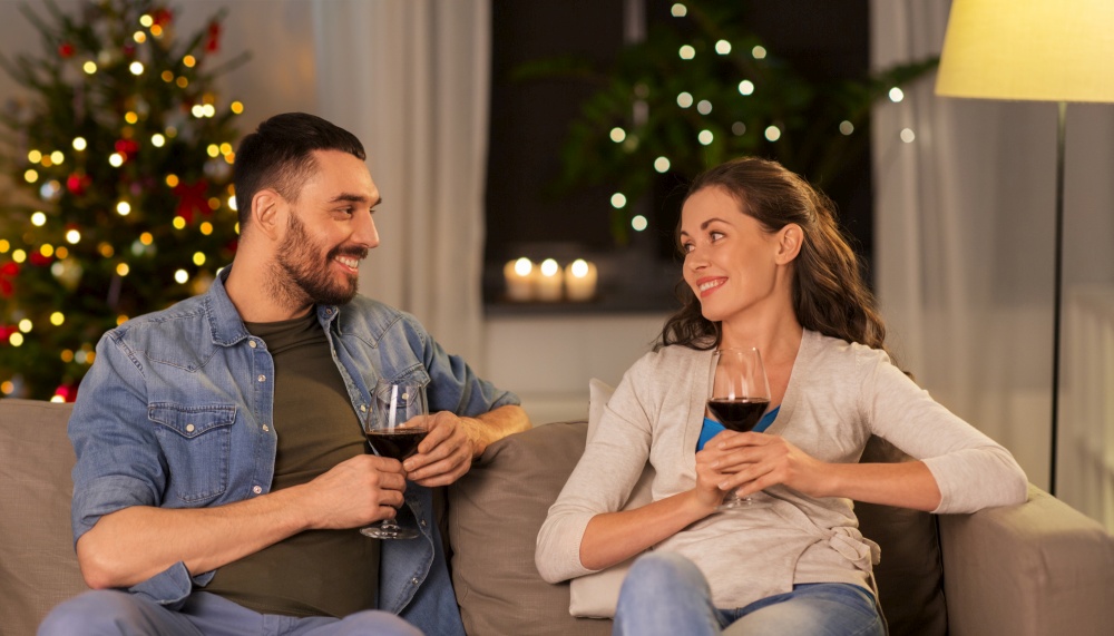 leisure, celebration and winter holidays concept - happy couple drinking red wine at home over christmas tree lights on background. happy couple drinking wine at home on christmas