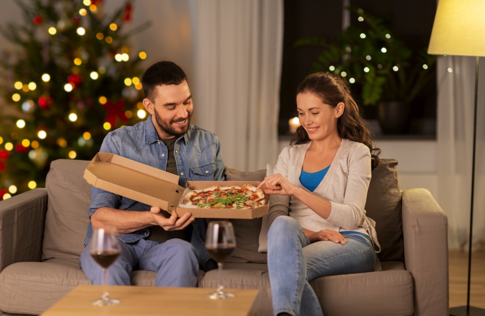 winter holidays, people and fast food concept - happy couple eating takeaway pizza at home in evening over christmas tree lights background. happy couple eating takeaway pizza on christmas