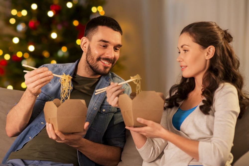 winter holidays, fast food and people concept - happy couple eating takeaway noodles with chopstick at home in evening over christmas tree lights background. happy couple eating takeaway noodles at home