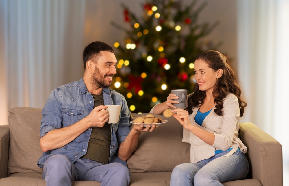 winter holidays, leisure and people concept - happy couple drinking tea or coffee with cookies at home over christmas tree lights on background. couple drinking tea with cookies on christmas