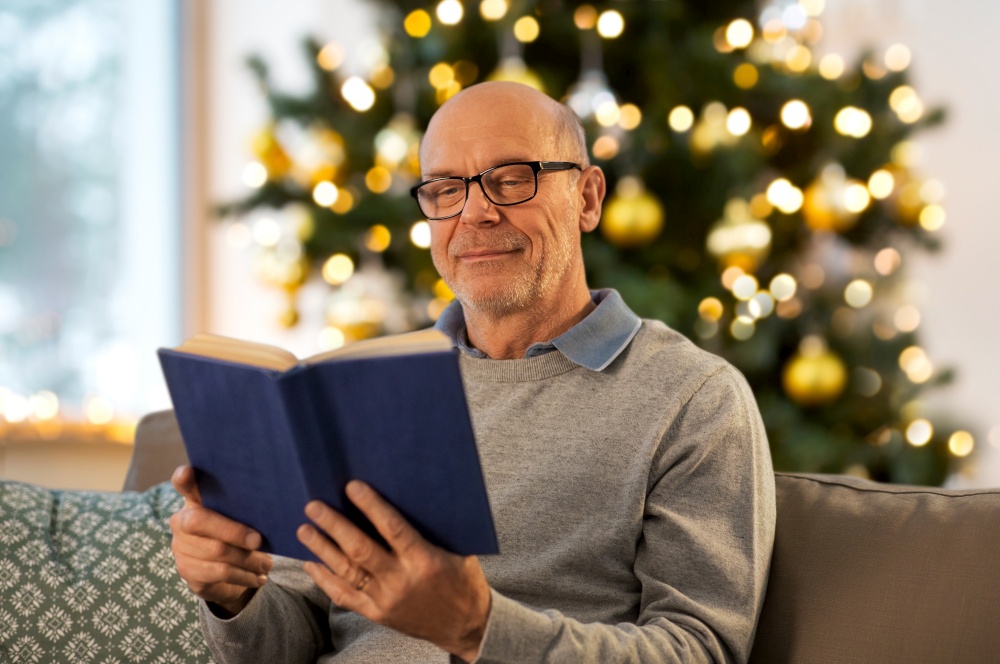 winter holidays, leisure and people concept - happy bald senior man sitting on sofa and reading book at home in evening over christmas tree lights on background. happy senior man reading book at home on christmas