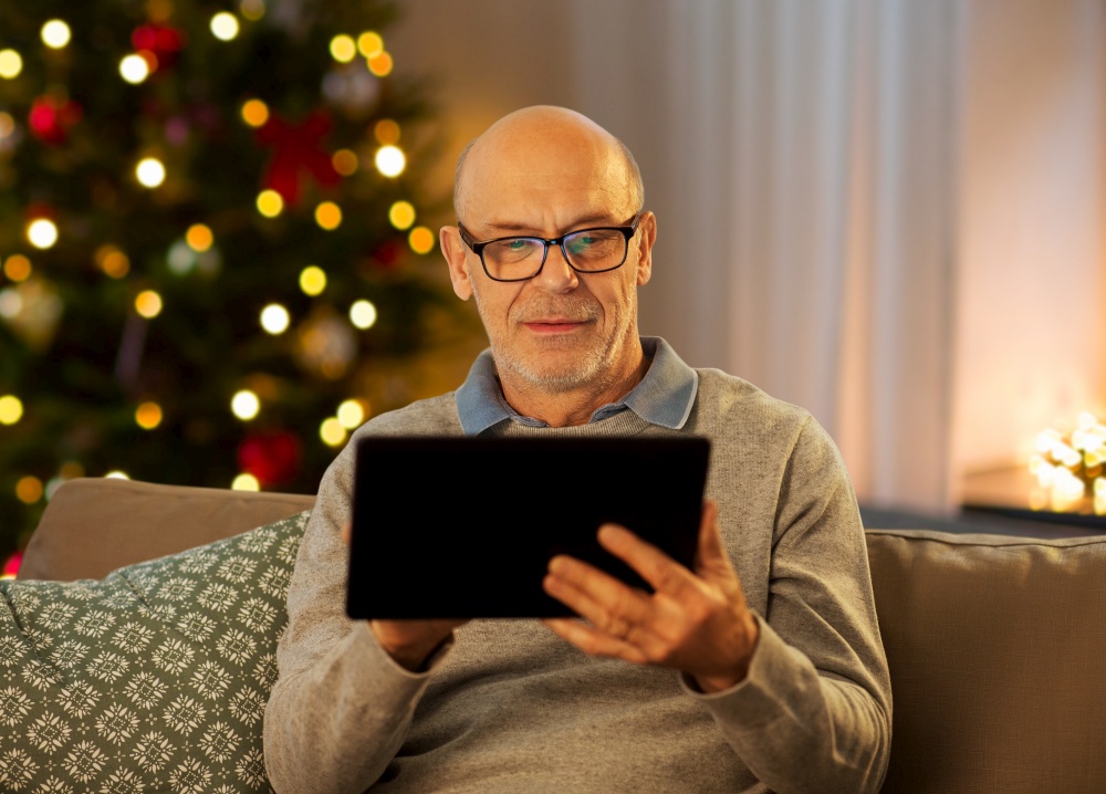 technology, winter holidays and people concept - happy bald senior man with tablet pc computer sitting on sofa at home in evening over christmas tree lights background. happy senior man with tablet computer at home