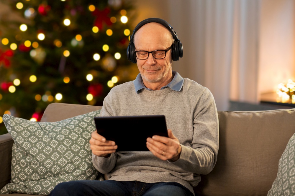 technology, winter holidays and people concept - happy senior man with tablet pc computer and headphones listening to music at home in evening over christmas tree lights background. senior man with tablet pc and headphones at home
