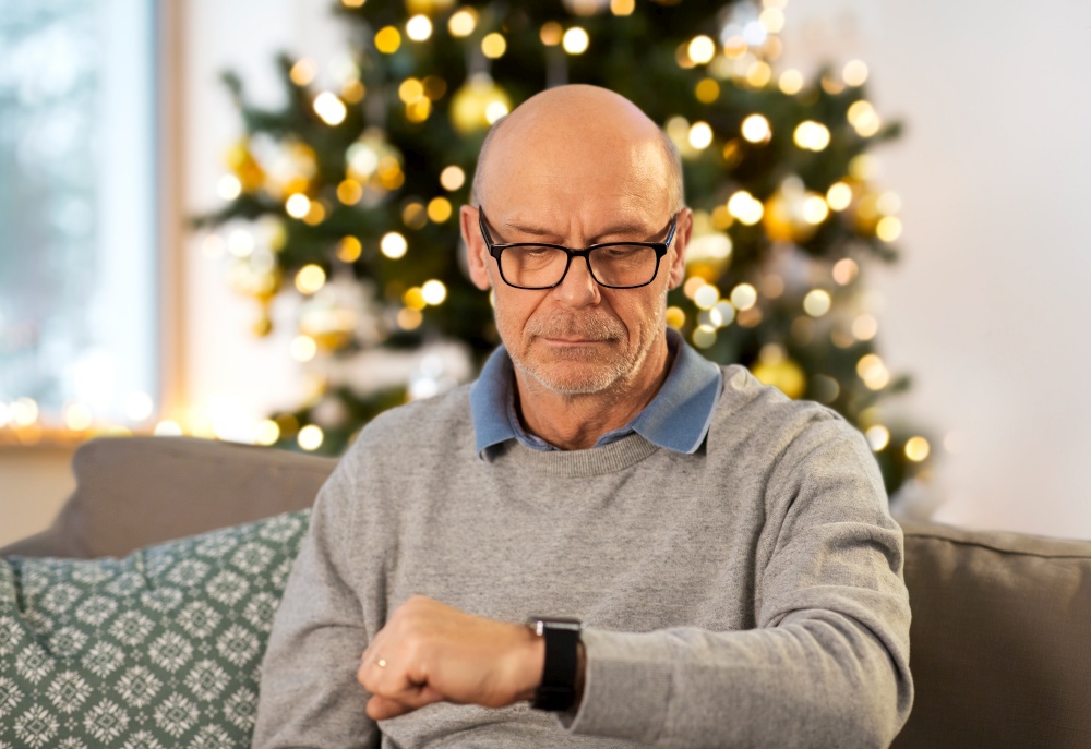 time, winter holidays and people concept - senior man looking at wristwatch at home in evening over christmas tree lights on background. senior man looking at watch at home on christmas