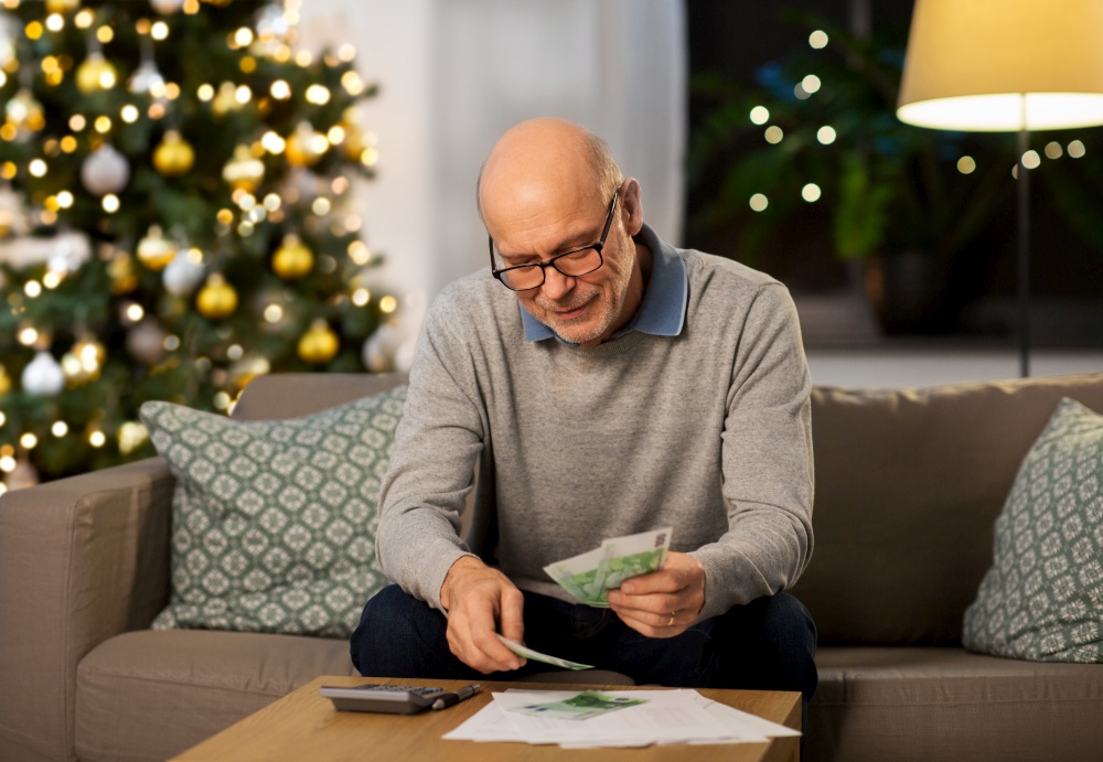 winter holidays, finances and people concept - smiling senior man with calculator and bills counting money at home in evening over christmas tree lights on background. senior man counting money at home on christmas
