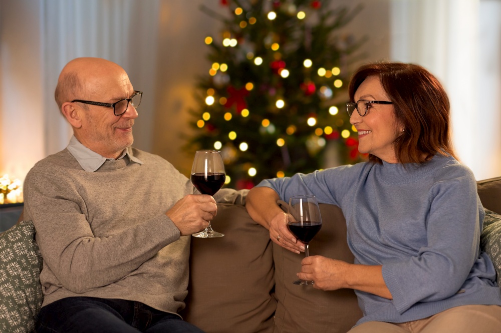 winter holidays, leisure and people concept - happy smiling senior couple toasting glasses of red wine at home in evening over christmas tree lights on background. happy senior couple with red wine on christmas