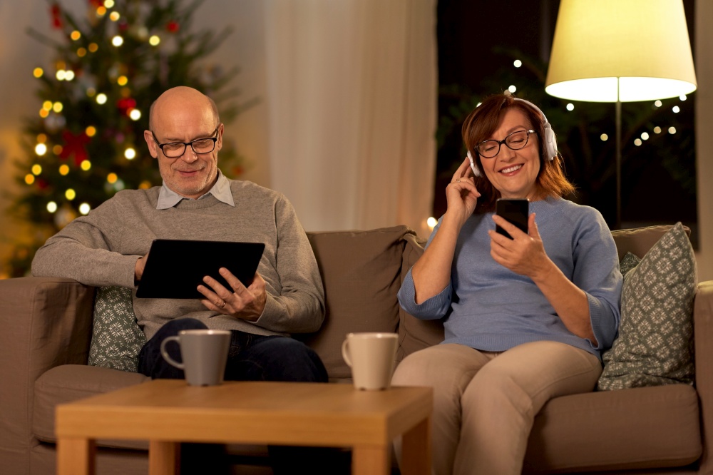 technology, winter holidays and people concept - happy senior couple with tablet pc computer, headphones and smartphone at home in evening over christmas tree lights on background. senior couple with gadgets at home on christmas