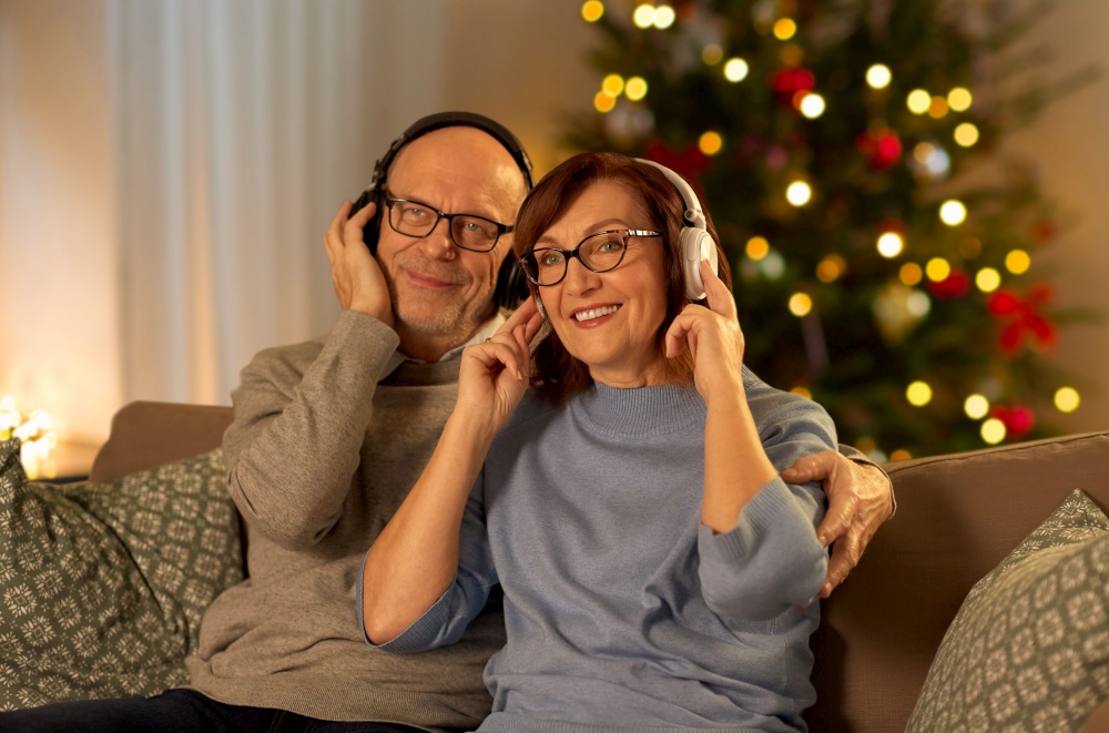 winter holidays, technology and people concept - happy senior couple with headphones listening to music at home in evening over christmas tree lights on background. senior couple with headphones on christmas