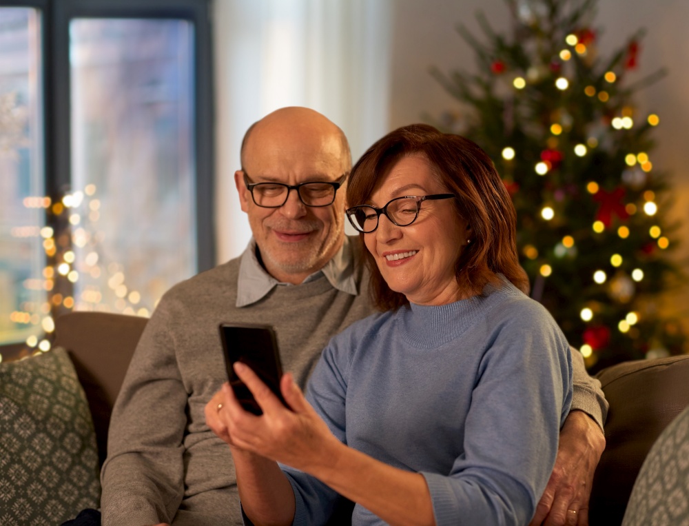 winter holidays, technology and people concept - happy senior couple with smartphone at home in evening over christmas tree lights on background. happy senior couple with smartphone on christmas