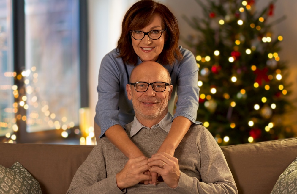 relationships, winter holidays and people concept - happy smiling senior couple hugging on sofa at home in evening over christmas tree lights on background. happy senior couple hugging at home on christmas