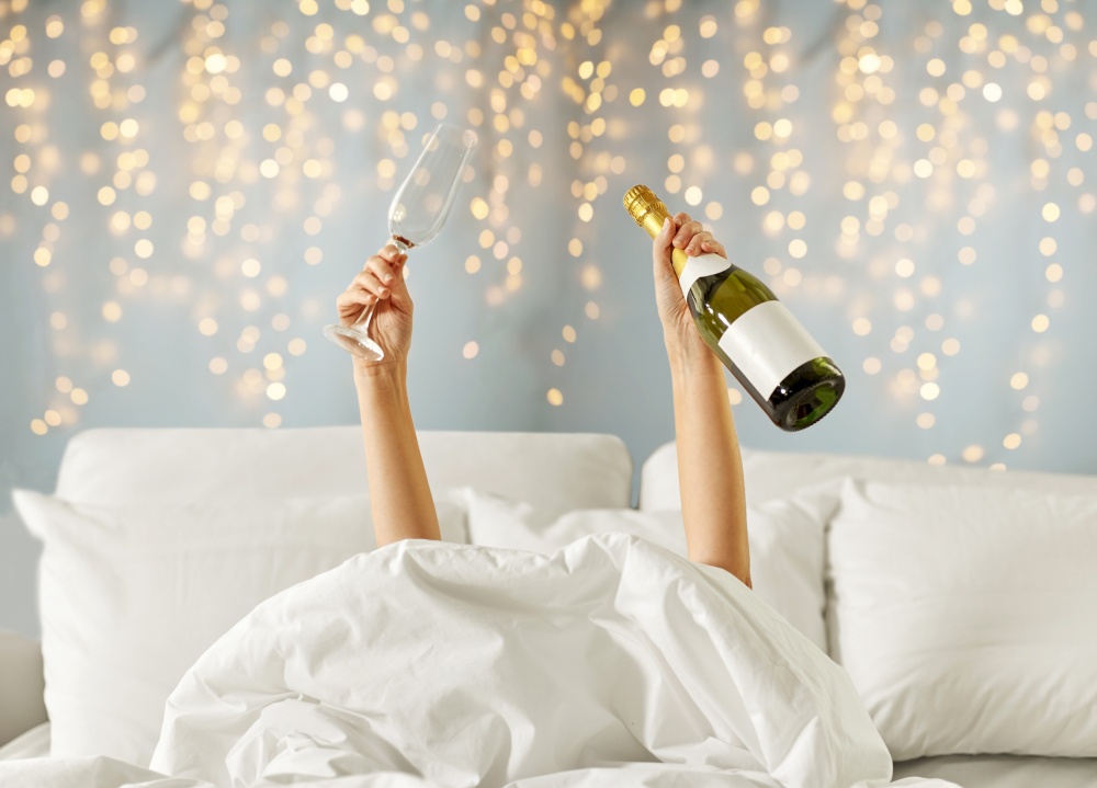 alcohol, comfort and morning concept - hands of young woman lying in bed with champagne glass and bottle over festive lights on background. hands of woman lying in bed with champagne