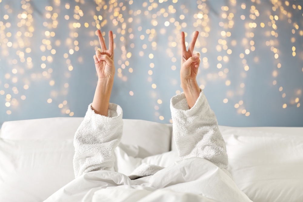 gesture, comfort and morning concept - hands of young woman in hotel robe lying in bed and showing peace hand sign over festive lights on background. hands of woman lying in bed and showing peace